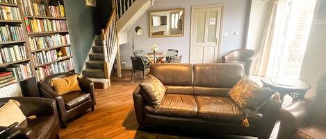 A living room with a leather sofa, armchairs, a bookshelf, a staircase, and a dining area.