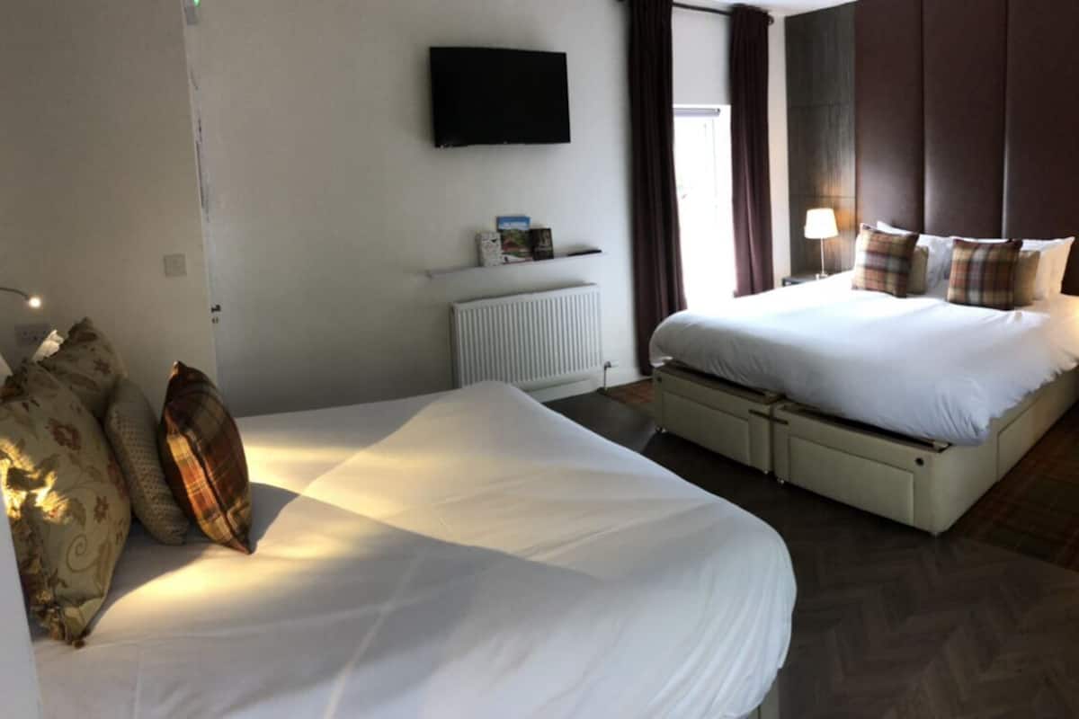 A hotel room with a large bed, a smaller bed, a TV mounted on the wall, a radiator, and a window with curtains.