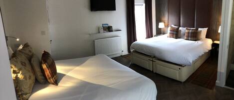 A hotel room with a large bed, a smaller bed, a TV mounted on the wall, a radiator, and a window with curtains.