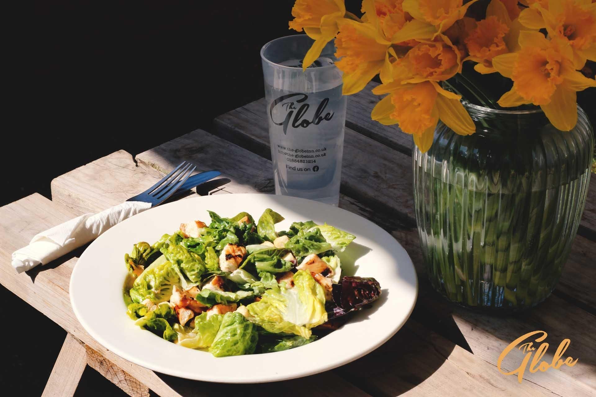 En vit tallrik med en sallad, ett glas med logotypen för ”The Globe” och en vas med gula blommor på ett träbord.