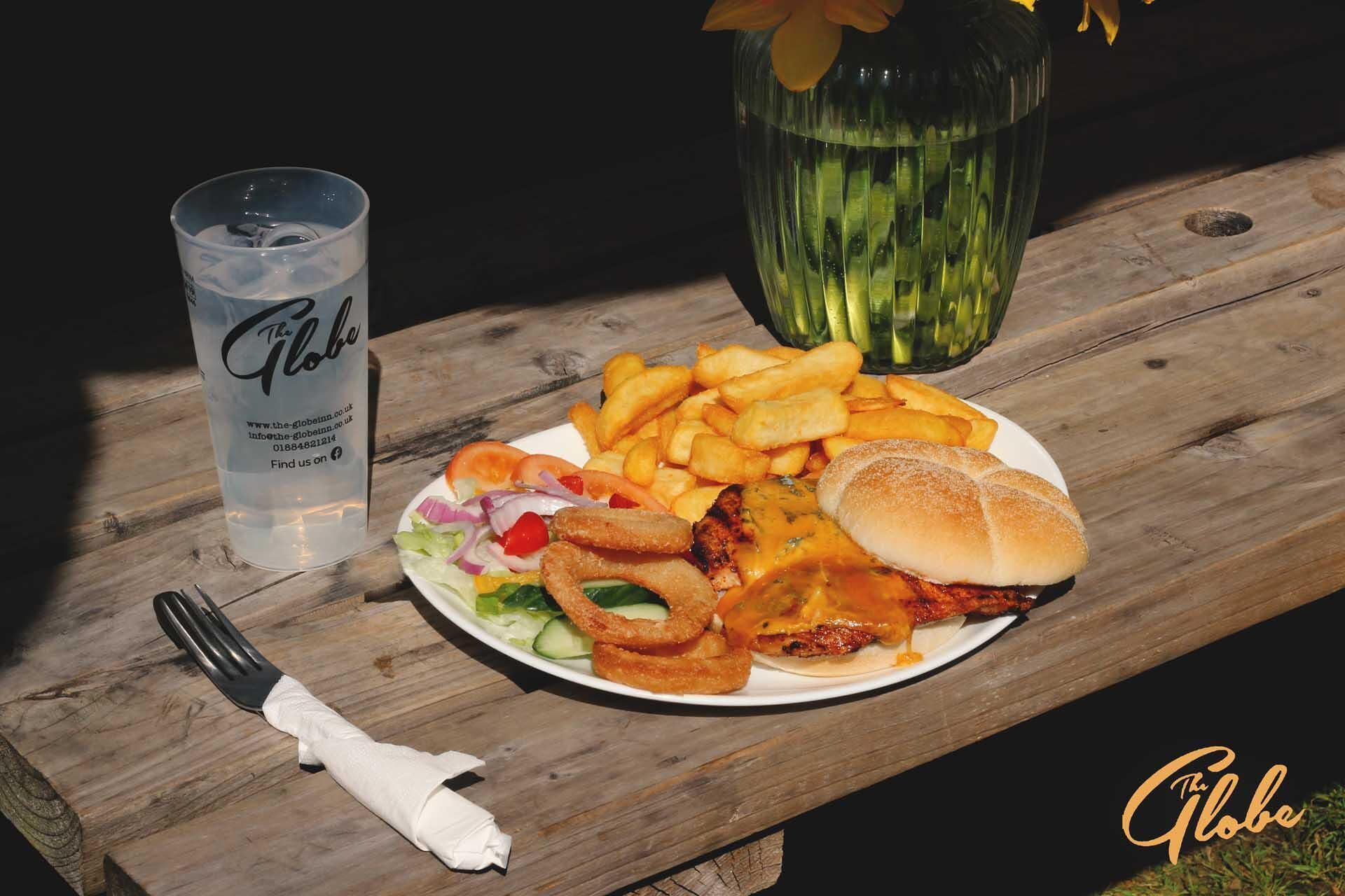 En tallerken mad bestående af en burger, pommes frites og løgringe, et glas vand og en serviet på et træbord.