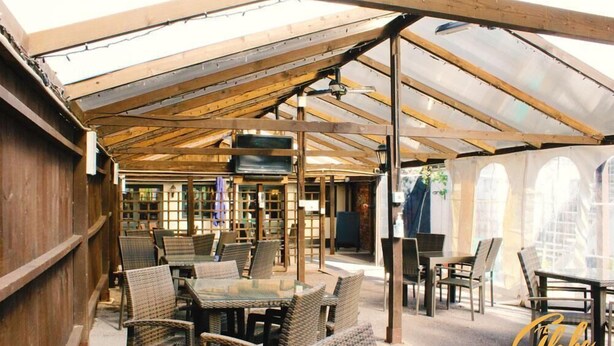 An outdoor patio with wooden tables and wicker chairs, a covered area with a metal roof, and a view of a building with large windows. - The Globe Inn (Tiverton)