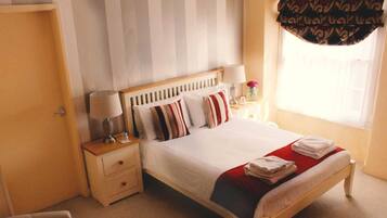 Una cama bien hecha con almohadas a rayas, una mesita de noche con lámpara y una ventana con cortinas.