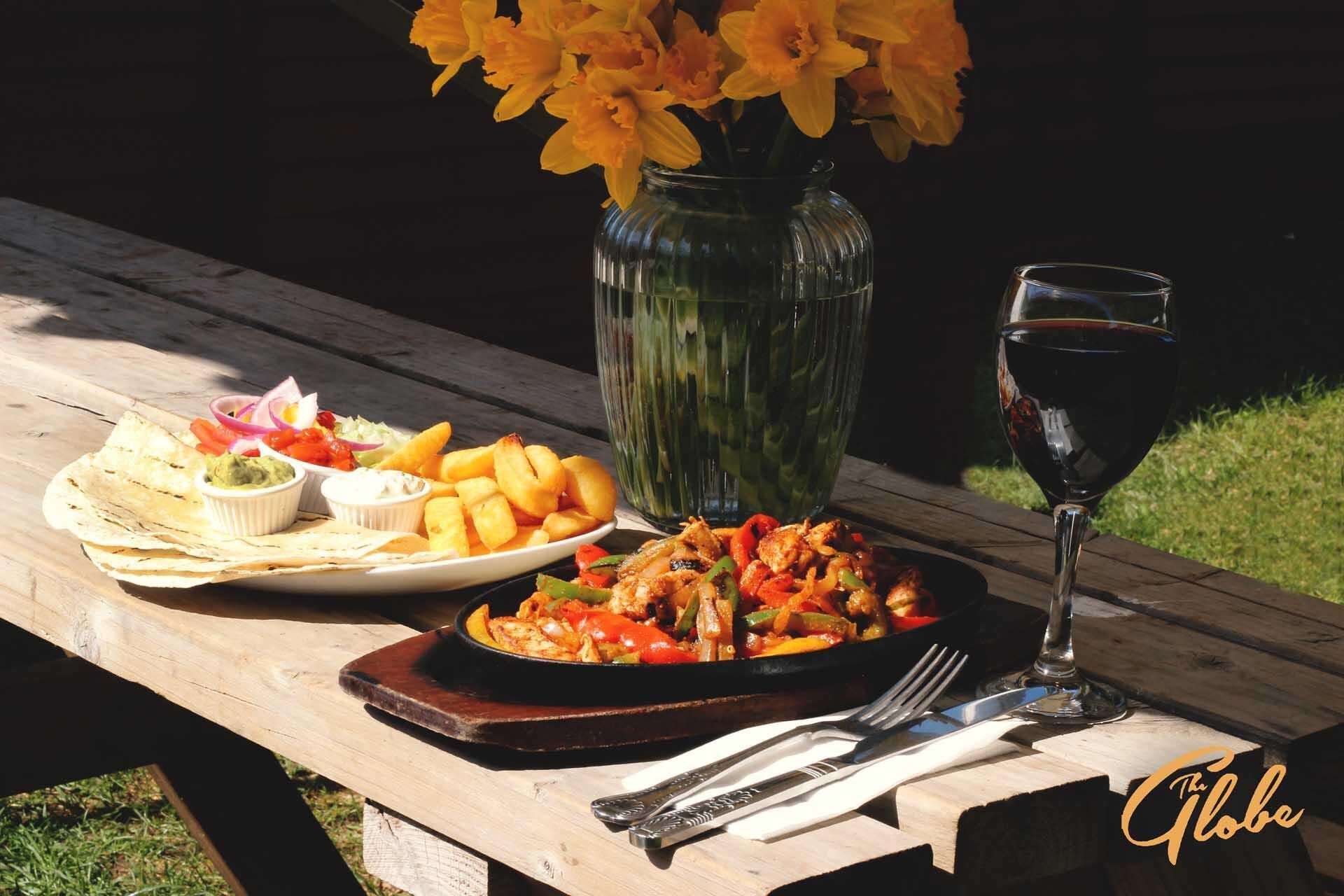 Un montaje para picnic con un plato de comida, una copa de vino tinto y un jarrón de flores amarillas sobre una mesa de madera.