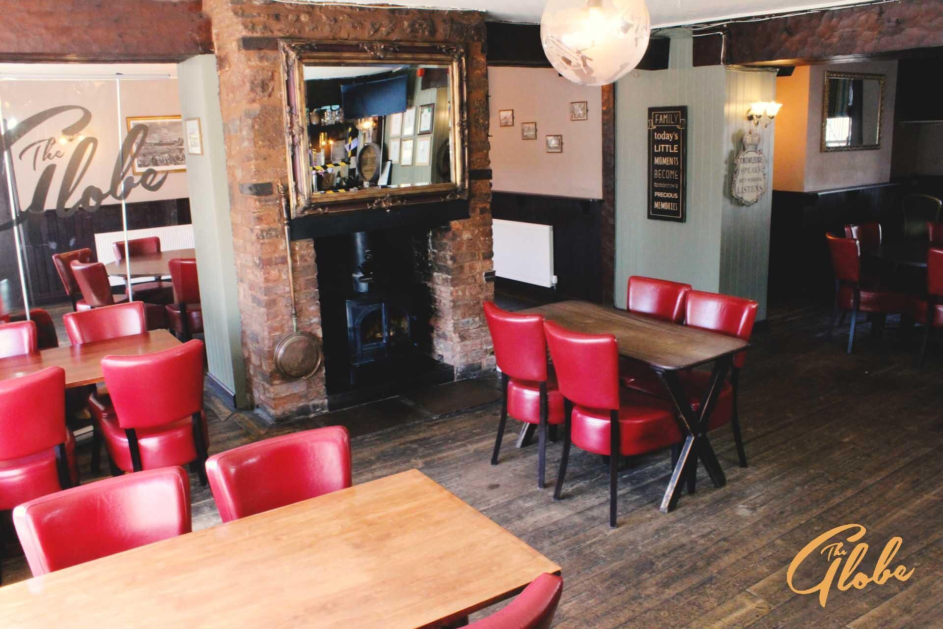 A restaurant interior with red chairs, wooden tables, a brick wall, a fireplace, and a mirror.