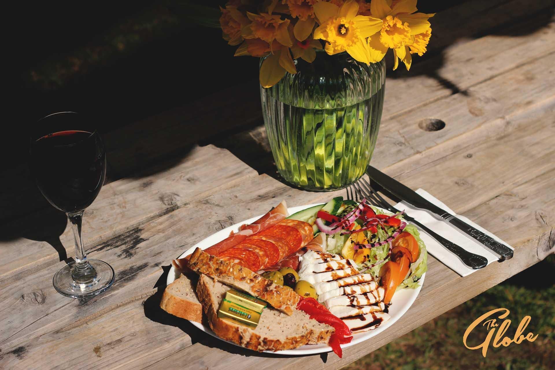 A wooden table with a plate of food, a glass of red wine, and a vase with yellow flowers.