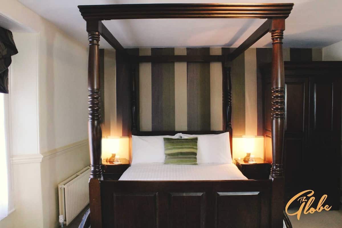 A four-poster bed with a white mattress and green pillows, flanked by two bedside tables with lamps, in a room with striped wallpaper and a radiator.