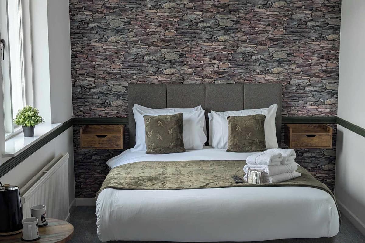 Una cama bien hecha con una pared de piedra de fondo, una mesita de noche con una planta y una ventana con cortinas.