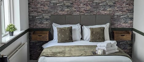 Ein ordentlich bezogenes Bett mit einer Steinwand im Hintergrund, ein Nachttisch mit Pflanze und ein Fenster mit Vorhängen.