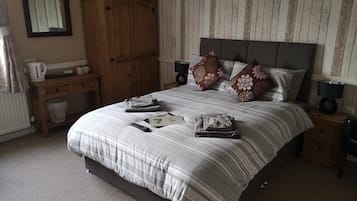 Un letto rifatto con cura, con copriletto a righe, cuscini e asciugamani piegati. Si vede un comodino in legno con una lampada e un tavolino con un bollitore e tazze. Sullo sfondo, un armadio in legno.