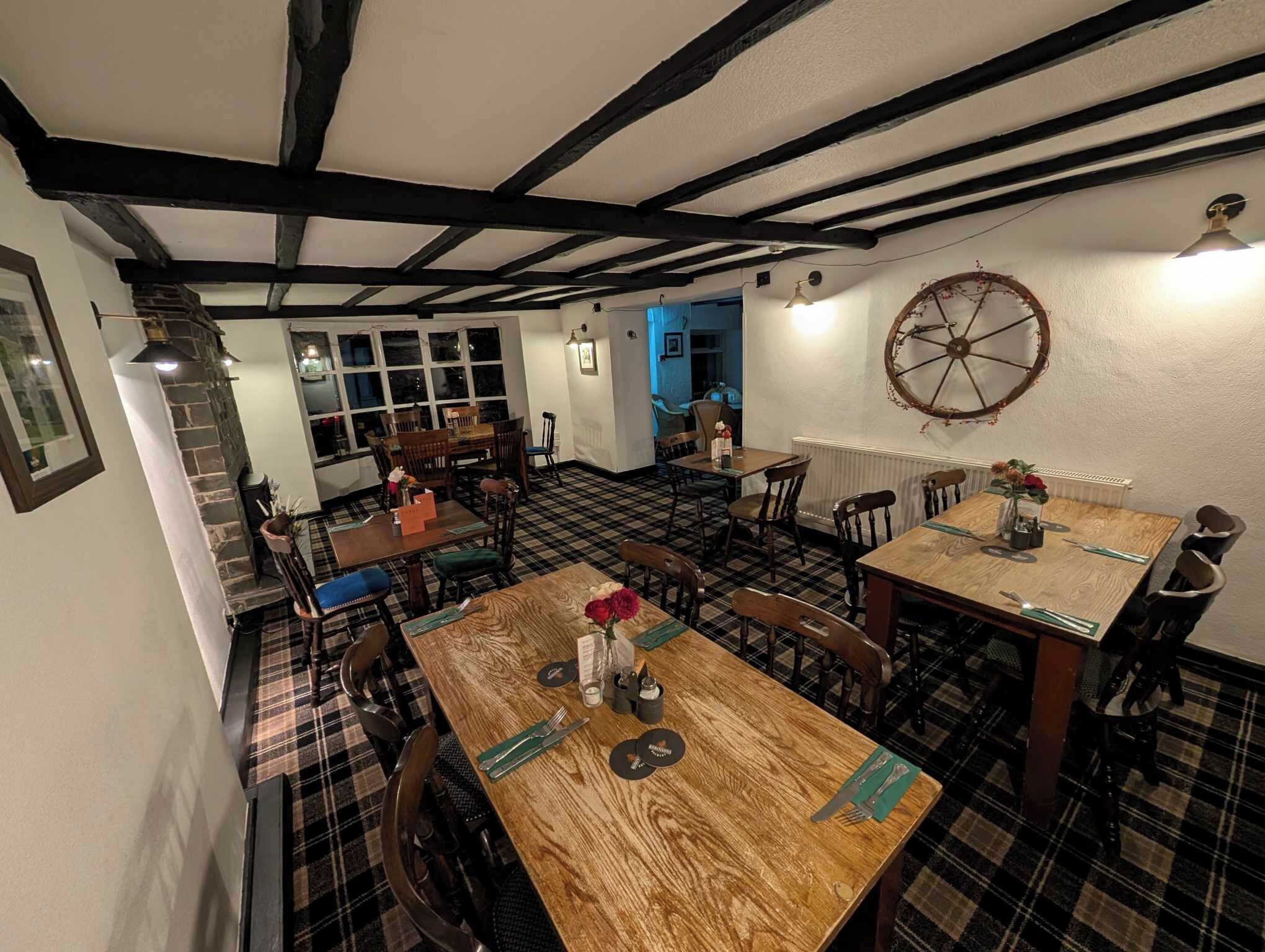Un espace repas avec un sol à carreaux, des tables et des chaises en bois. On y trouve une grande roue décorative au mur et une fenêtre avec des rideaux.
