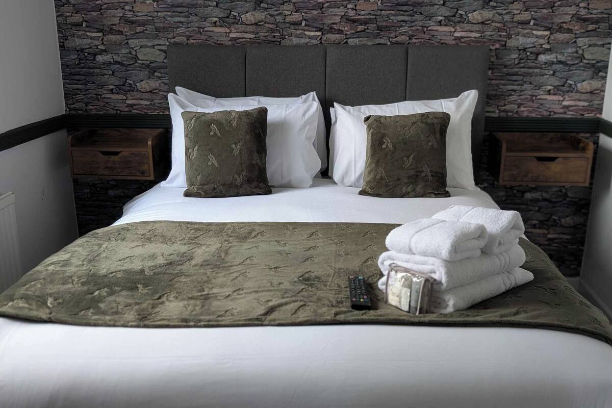 Una cama bien hecha con un cabecero gris, dos almohadas blancas, dos almohadas verdes, una manta gris y una pila de toallas blancas.