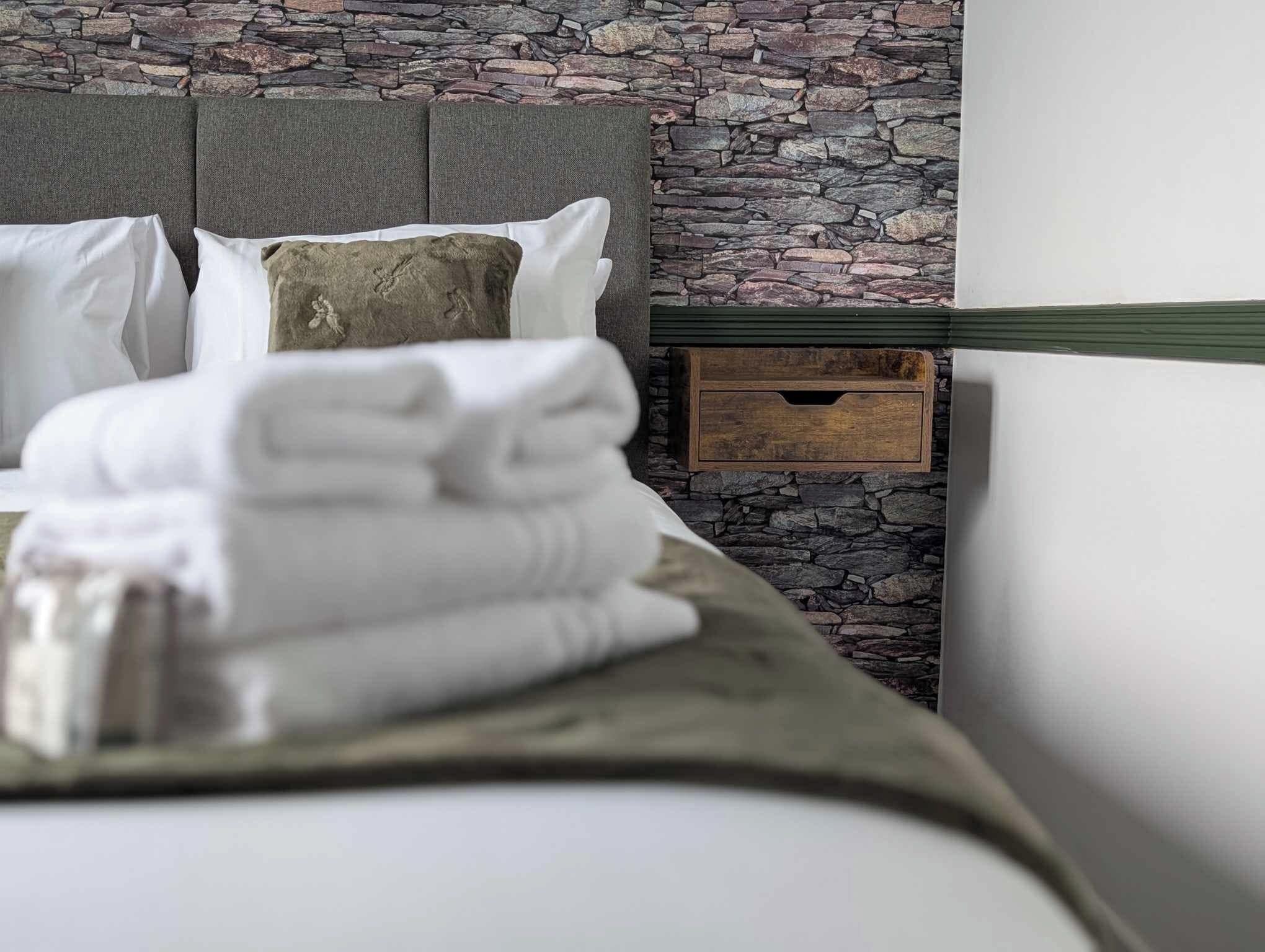 A neatly made bed with white and green linens, a stone wall, and a wooden nightstand with a drawer.