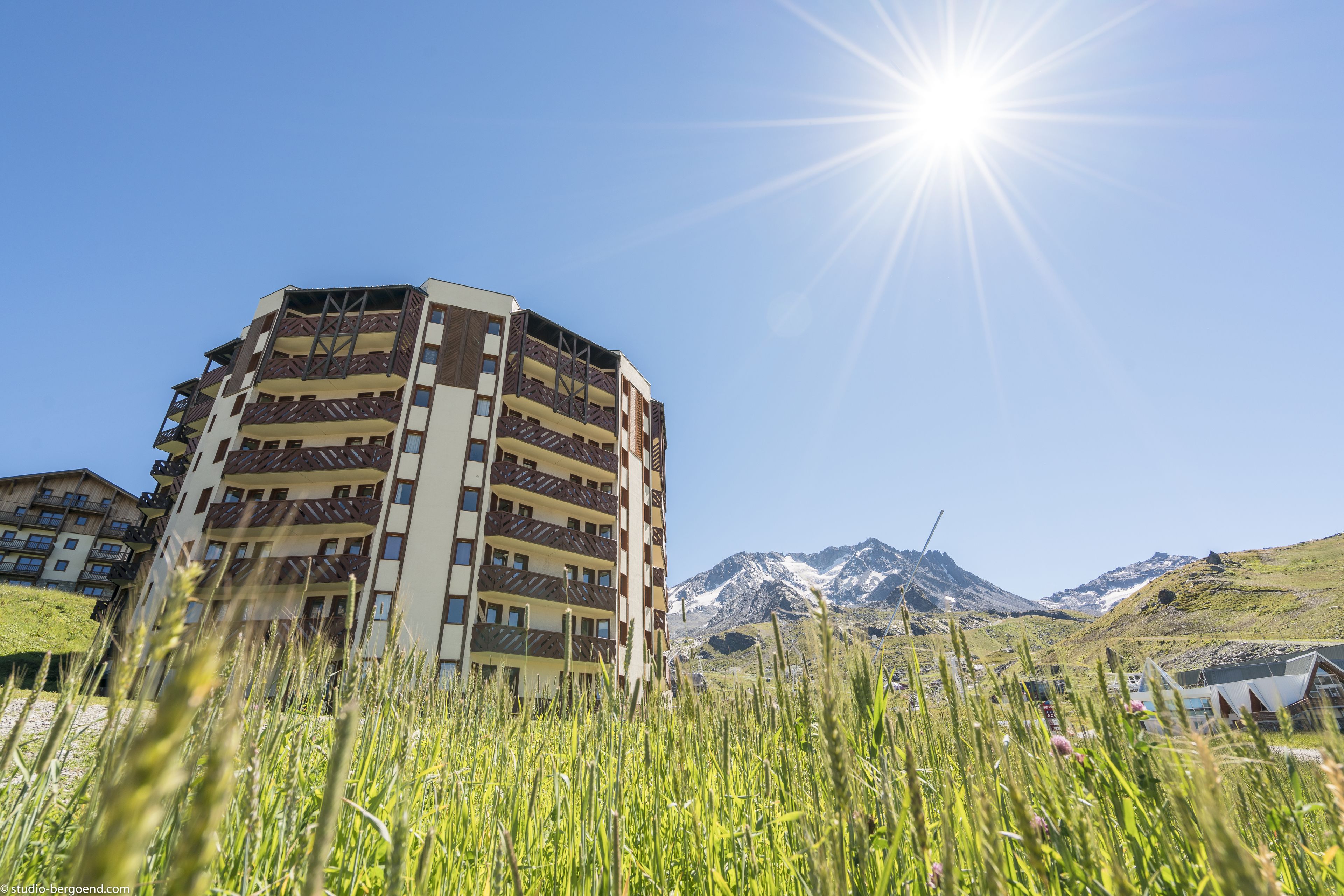 A multi-story building with balconies, set against a backdrop of mountains and clear sky.