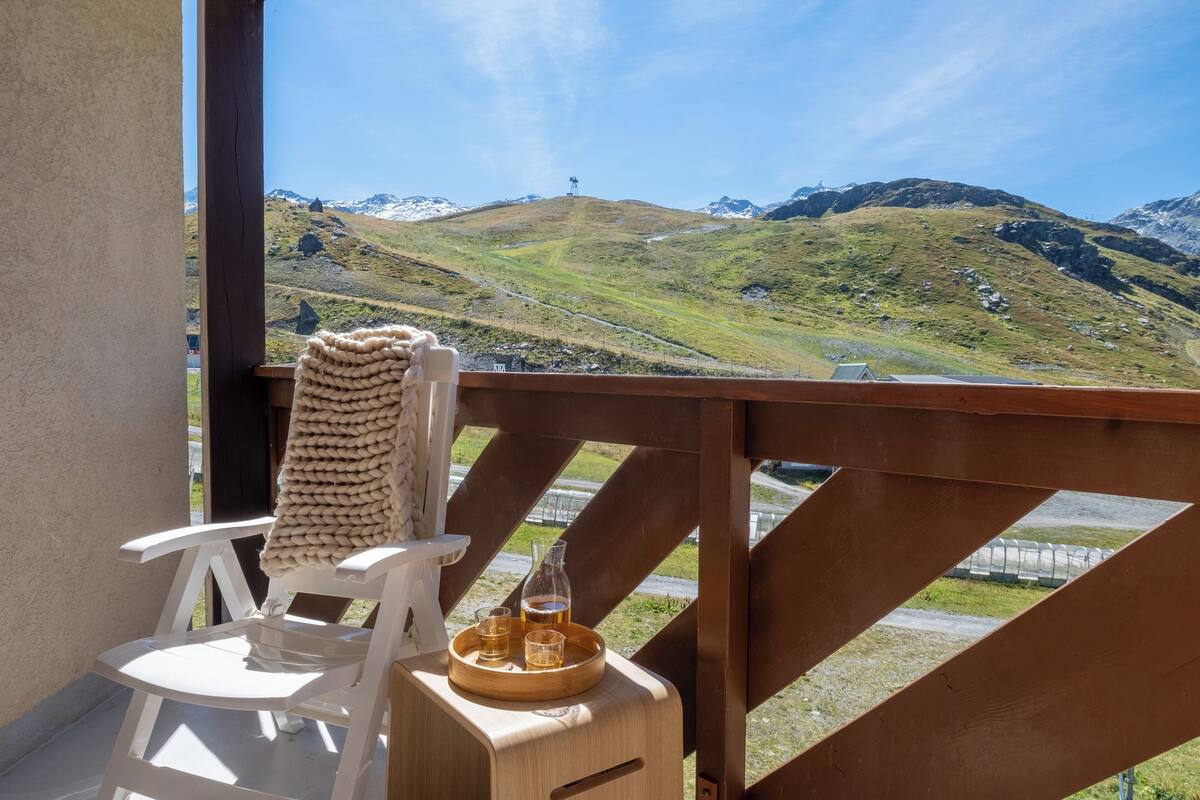 A balcony with a wooden railing, a white chair, a wooden table with a tray holding glasses and a bottle, and a view of green hills and mountains.