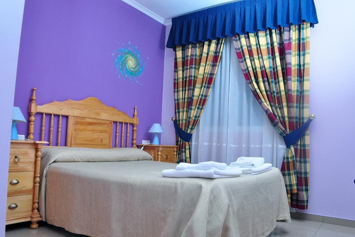 A bedroom with a wooden headboard, a bed with a beige bedspread, a nightstand with a lamp, and a window with plaid curtains.