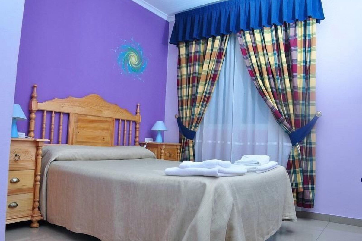 A bedroom with a bed, a wooden headboard, a nightstand, a lamp, and a window with plaid curtains.