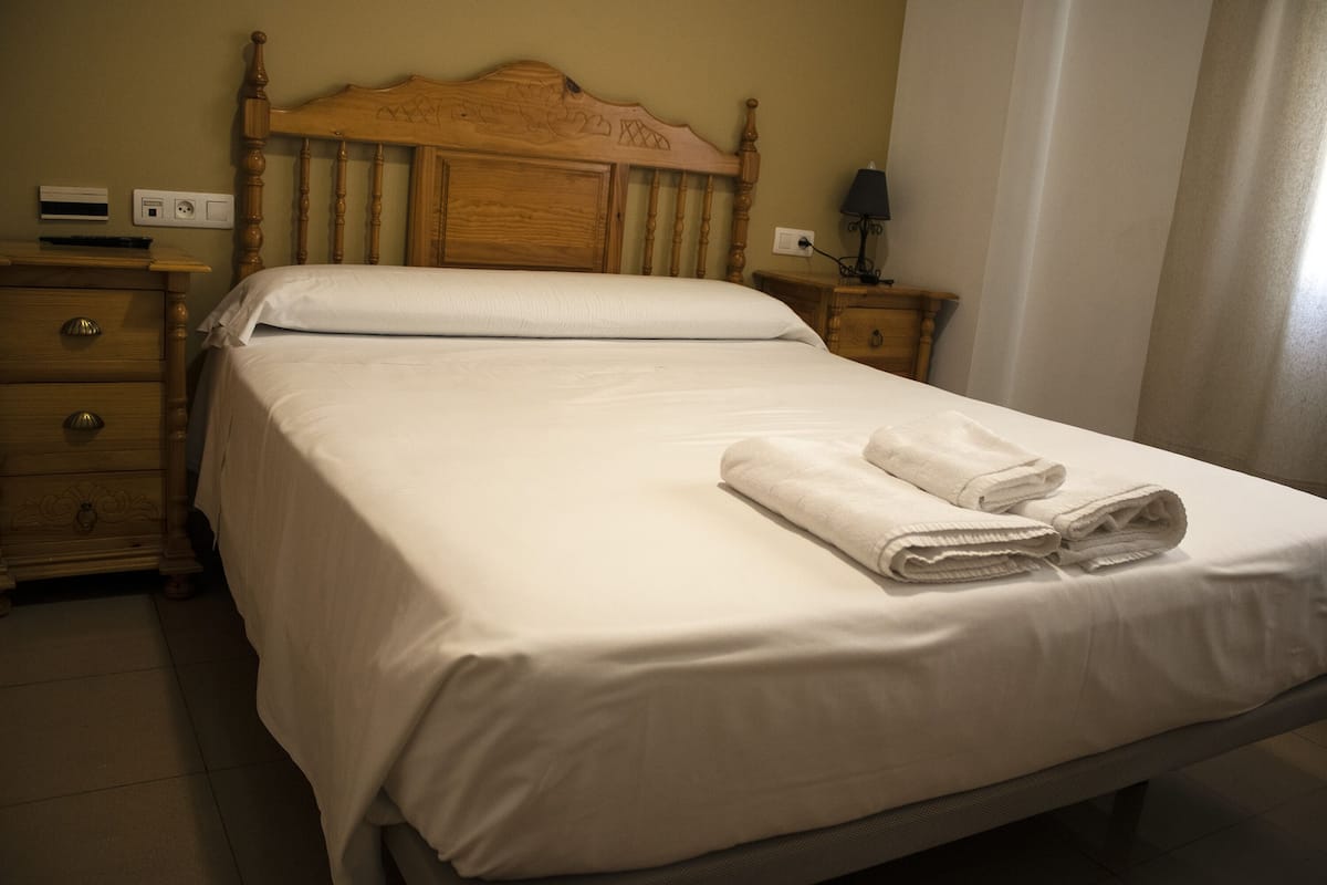 A neatly made bed with white linens and folded towels, a wooden headboard, a bedside table with a lamp, and a wall-mounted air conditioning unit.