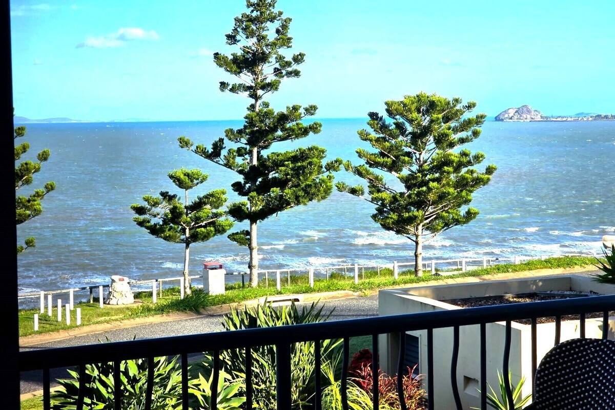 Un balcón con vistas al mar, con tres pinos y una barandilla.
