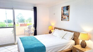 Een hotelkamer met een bed, twee nachtkastjes met lampen, een stoel, een balkon met uitzicht op groen en een schilderij aan de muur.