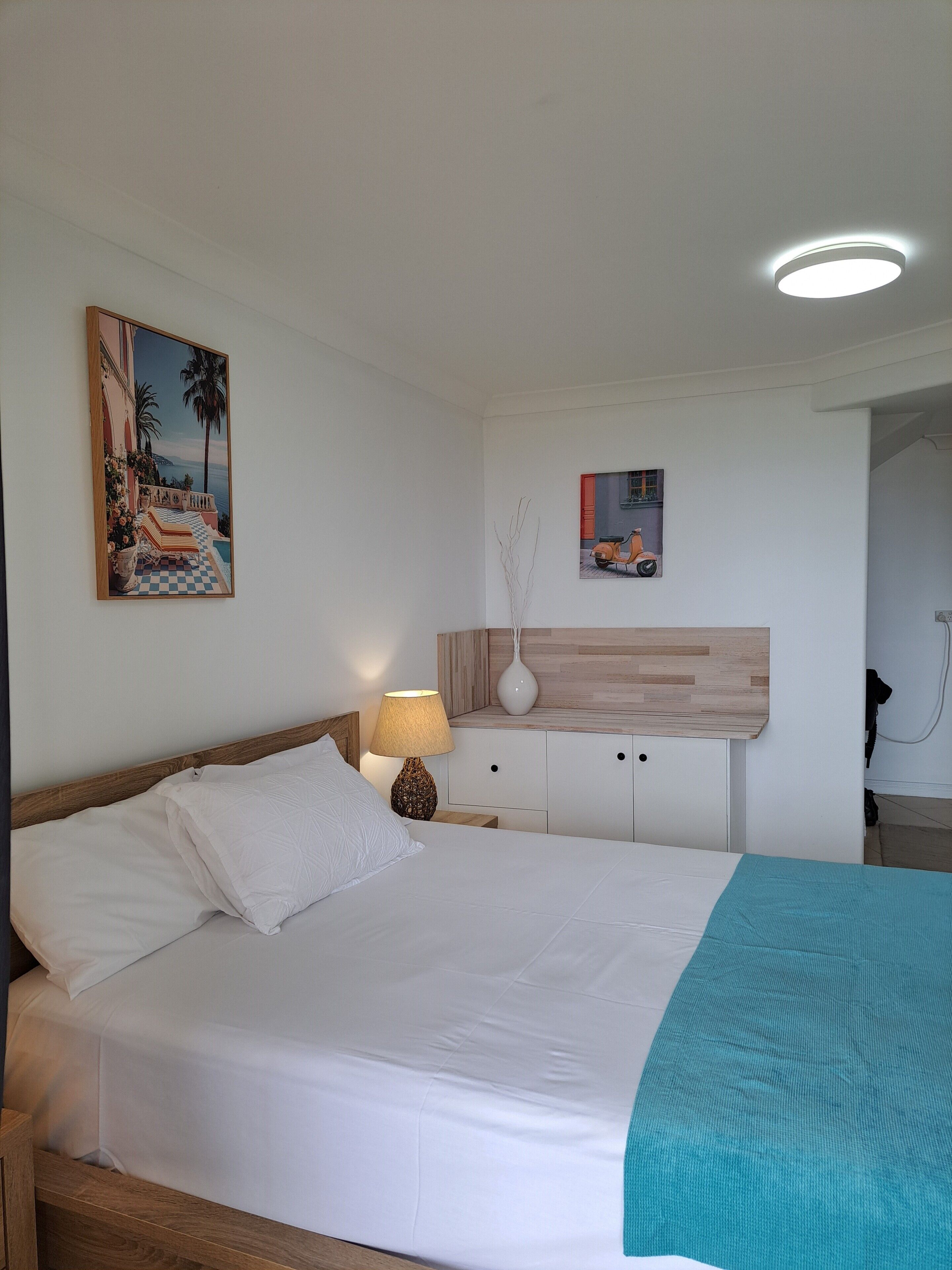 A hotel room with a bed, a nightstand, a lamp, a painting, a small cabinet, and a ceiling light.