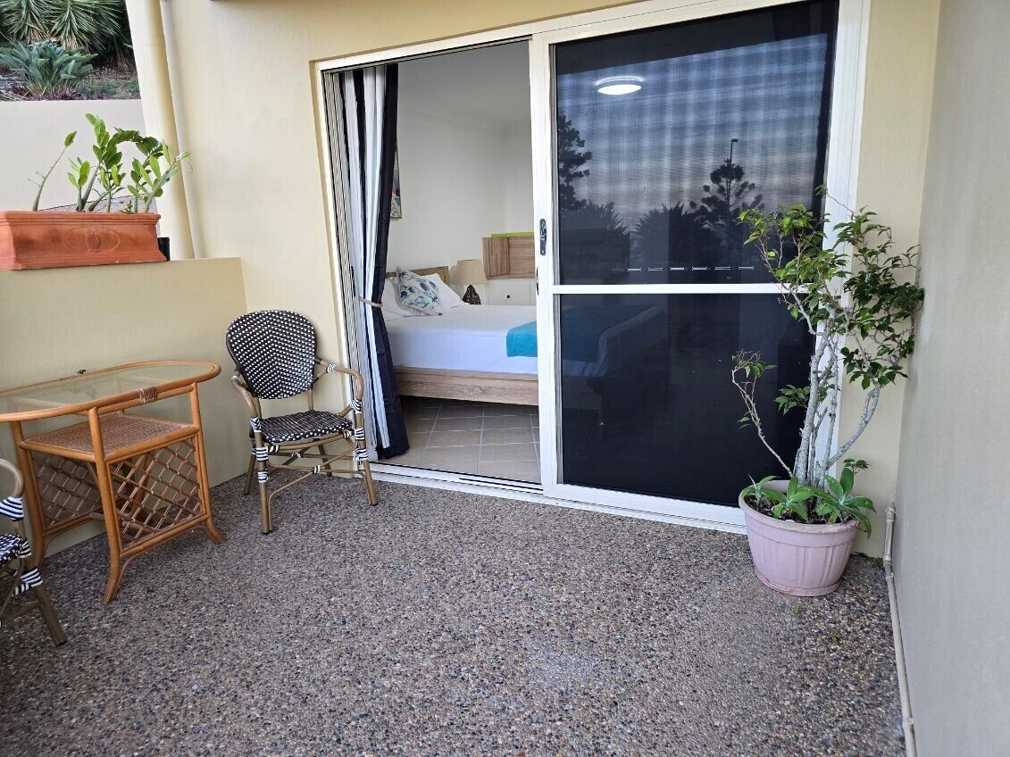A balcony with a table and chairs, a sliding glass door leading to a bedroom, and a potted plant.