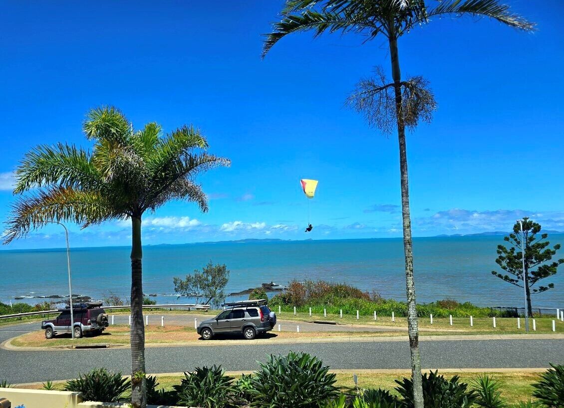 A coastal road with parked cars, palm trees, and a clear view of the sea and distant mountains.