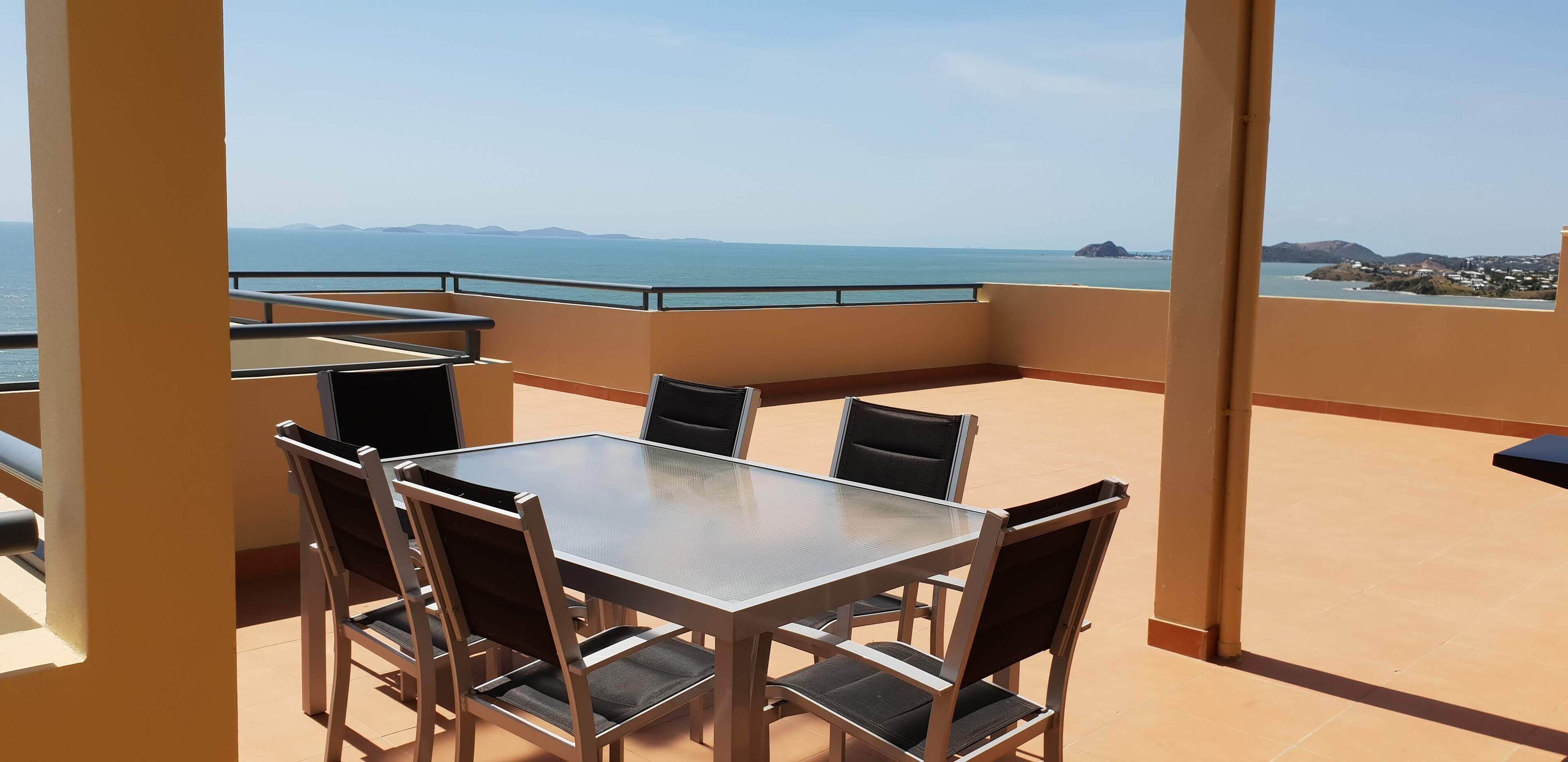 A balcony with a glass table and chairs, overlooking a coastal view with mountains in the distance.