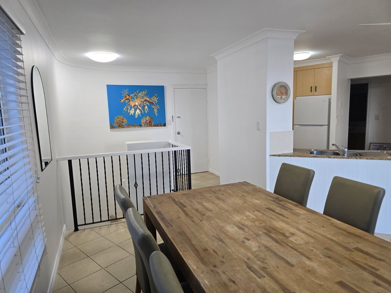 A dining area with a wooden table and chairs, a kitchen with white cabinets and a refrigerator, and a blue painting on the wall.
