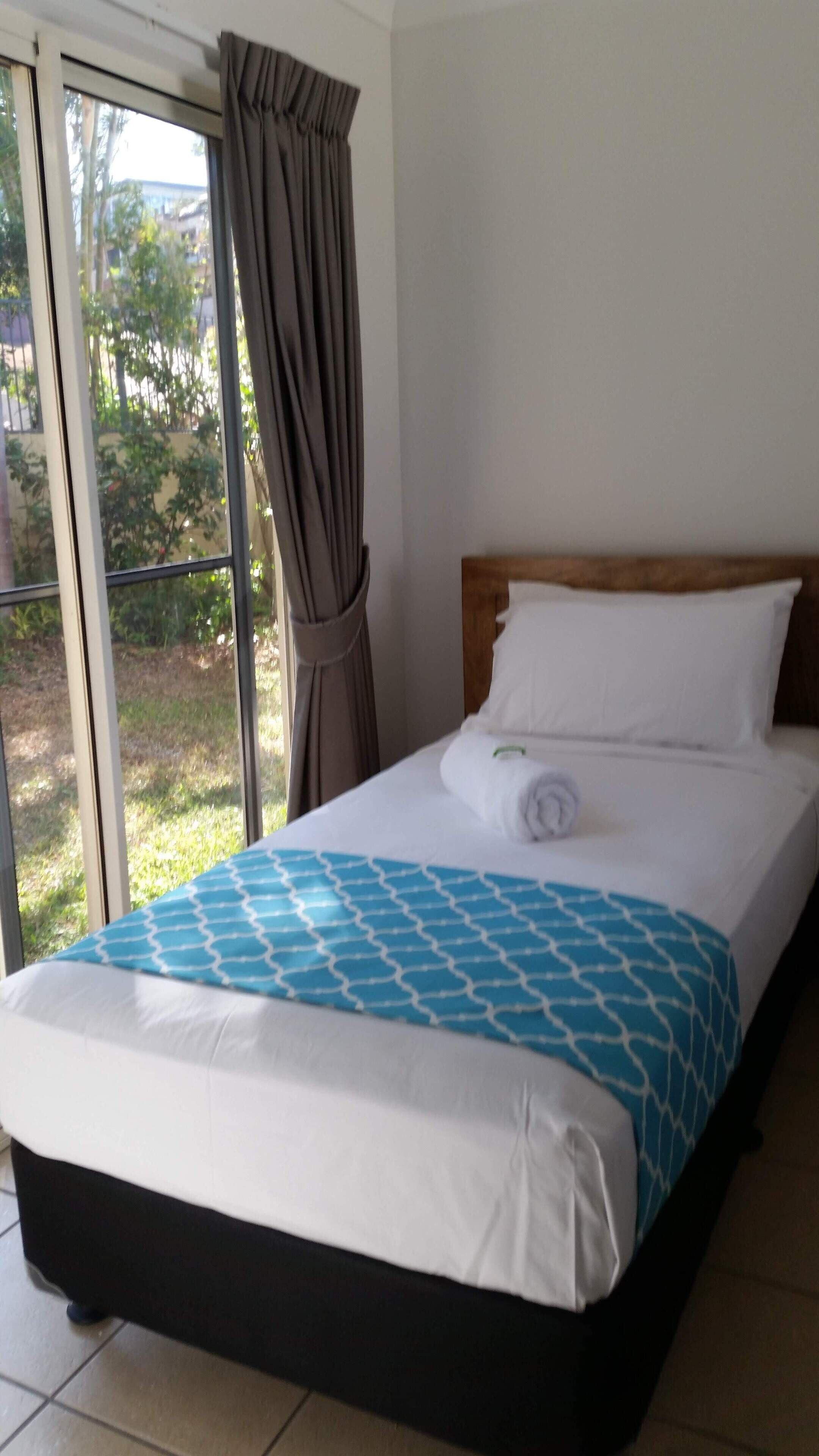 A hotel room with a bed, a window with curtains, and a view of greenery outside.