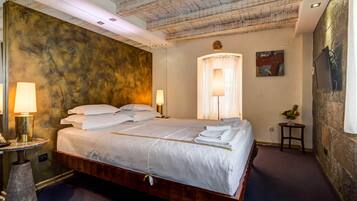 A neatly made bed with white linens, a round bedside table with a lamp, and a wall-mounted TV.