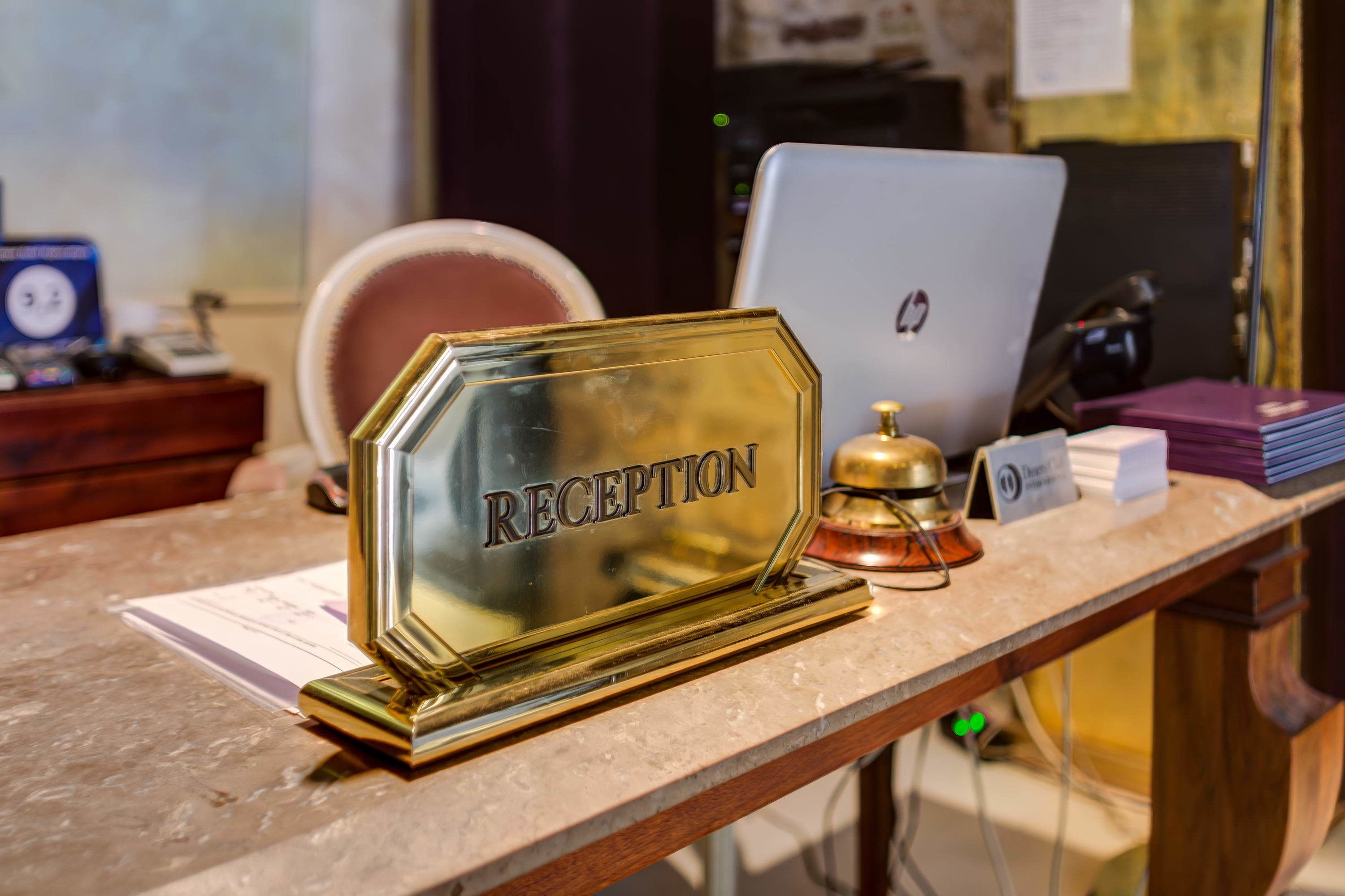 A reception desk with a glass nameplate labeled "RECEPTION," a laptop, a bell, and some documents.