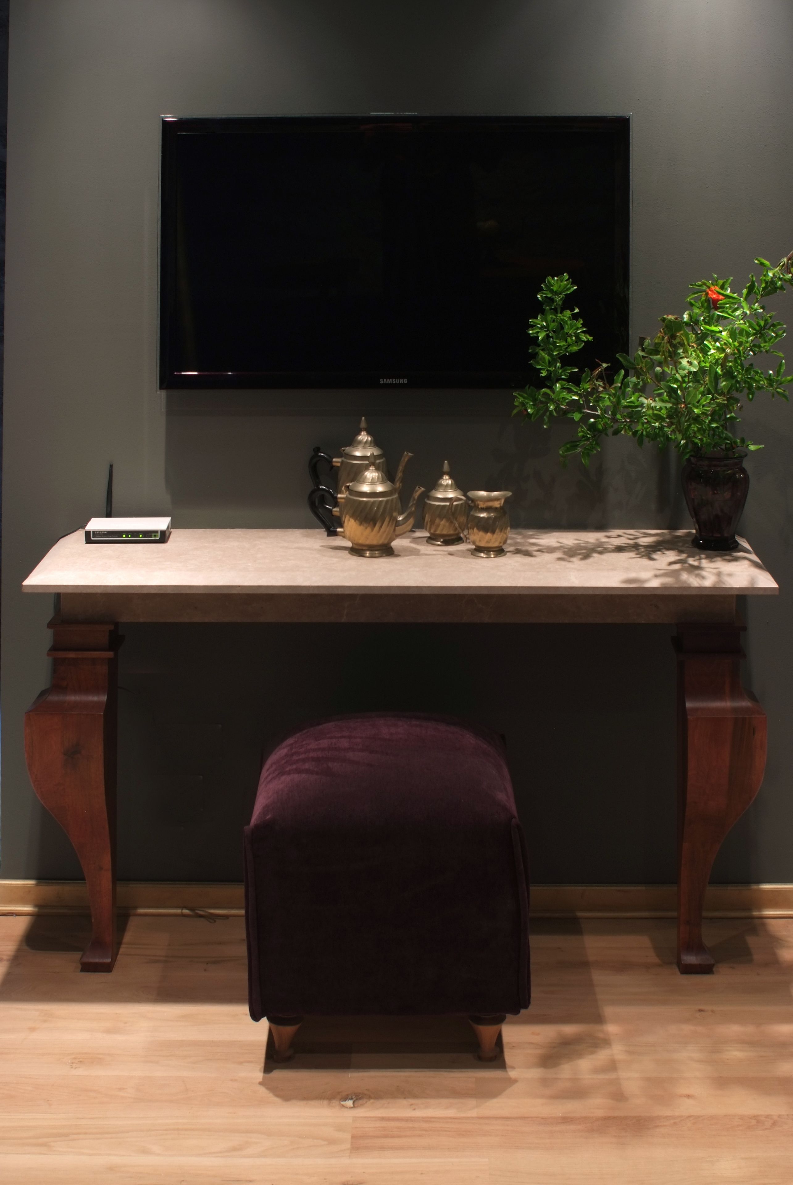 A room with a flat-screen TV mounted on the wall, a wooden table with a tea set, and a dark-colored ottoman.