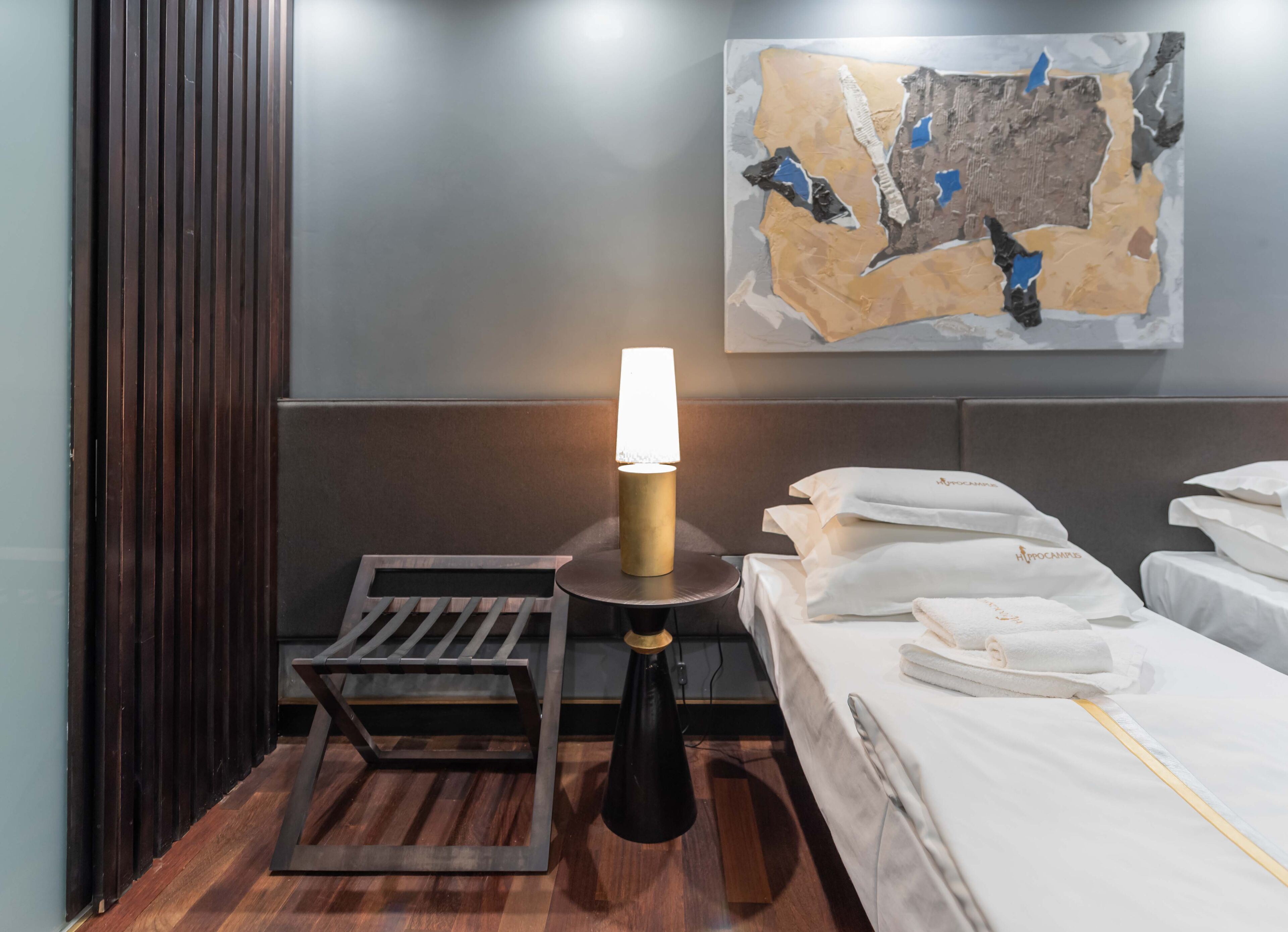 A hotel room with a bed, a small table with a lamp, a wooden bench, and a wall-mounted artwork.
