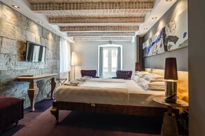 A hotel room with a stone wall, a bed with a wooden headboard, a bench, a table, and a lamp. - Hotel Hippocampus (Kotor)