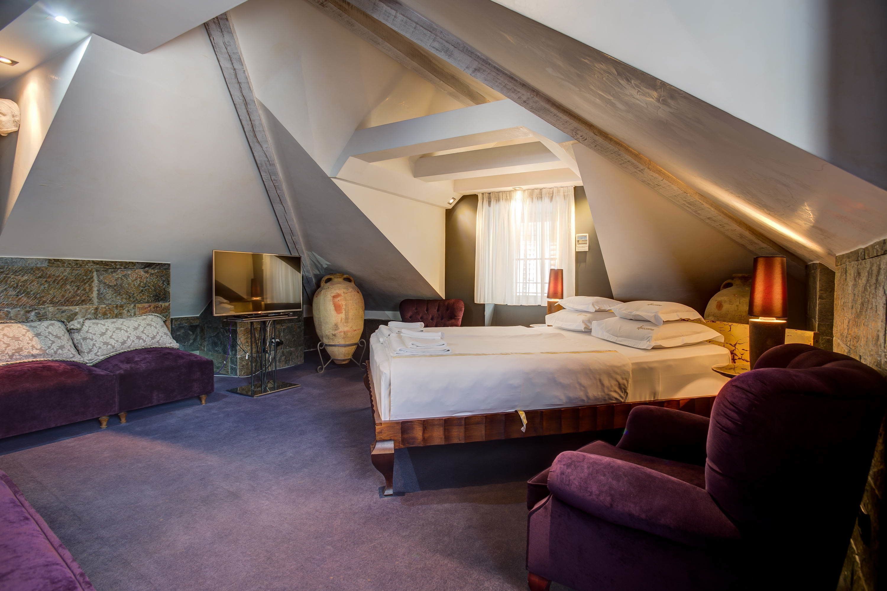A high-ceilinged room with a sloped ceiling, a bed with white linens, a large vase, a wooden cabinet, and a purple armchair.