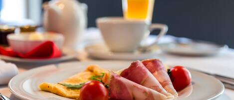 Un plato de desayuno con jamón y tomates, un vaso de zumo de naranja y una tetera al fondo.