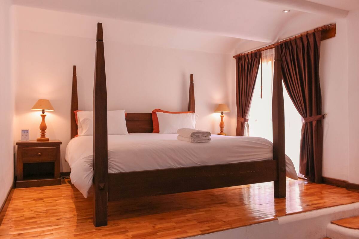 Una cama con dosel, ropa de cama blanca, cabecera de madera y dos mesitas de noche con lámparas.