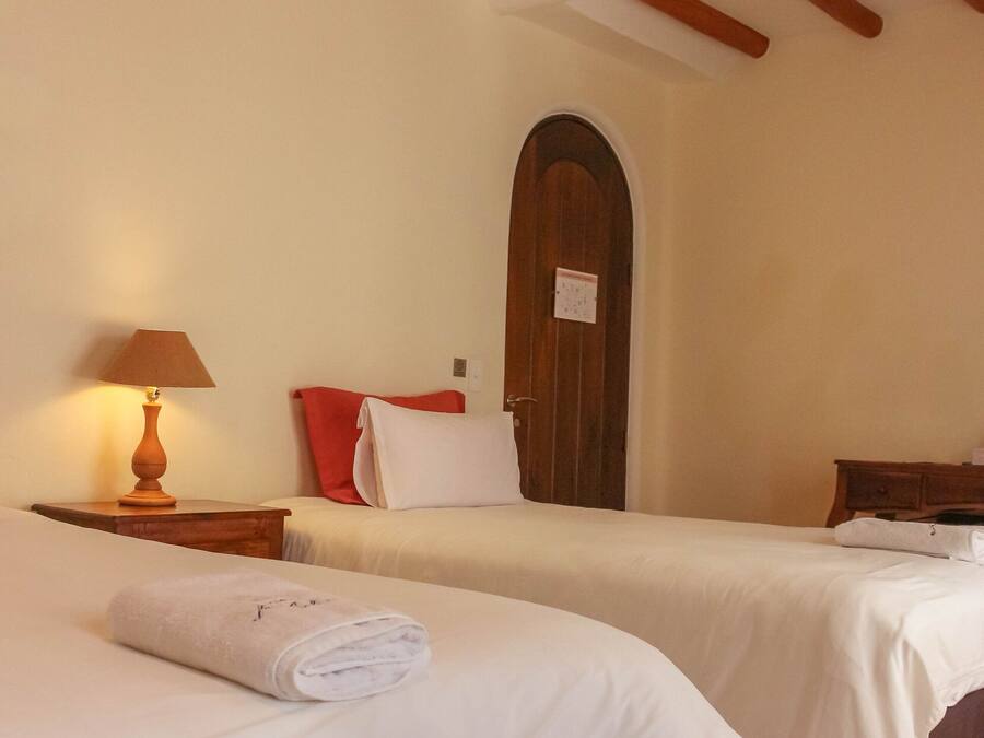 A hotel room with two beds, wooden beams on the ceiling, a wooden door, and a nightstand with a lamp.