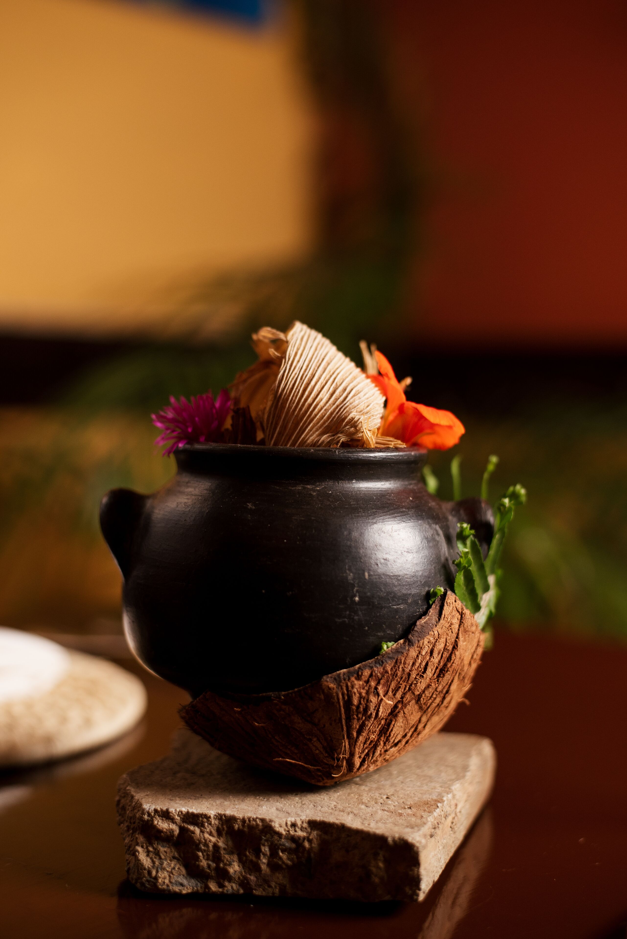 Een zwarte keramieken pot op een houten blok, samen met een geweven mand en gedroogde bloemen.