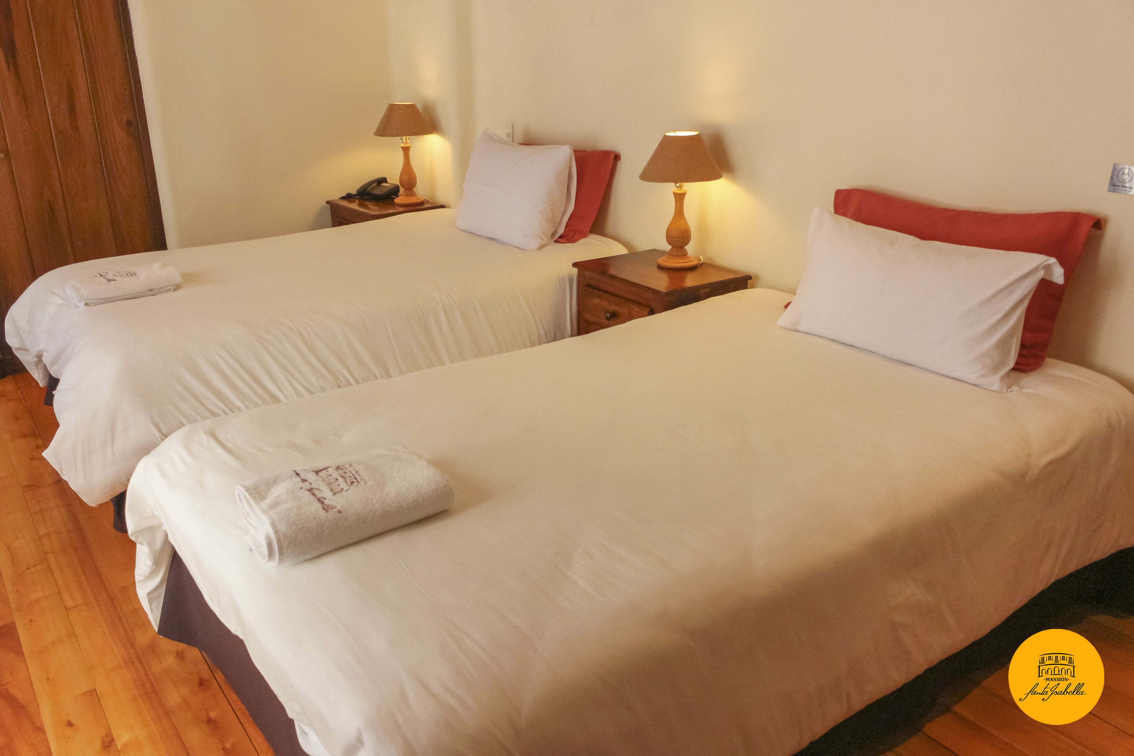 A hotel room with two beds, each with white linens and red pillows, wooden nightstands with lamps, and a wooden floor.