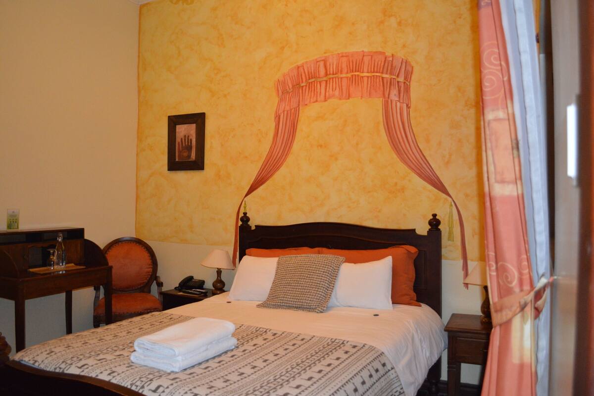 Habitación de hotel con cama con dosel, mesita de noche con lámpara, una silla e imagen enmarcada en la pared.