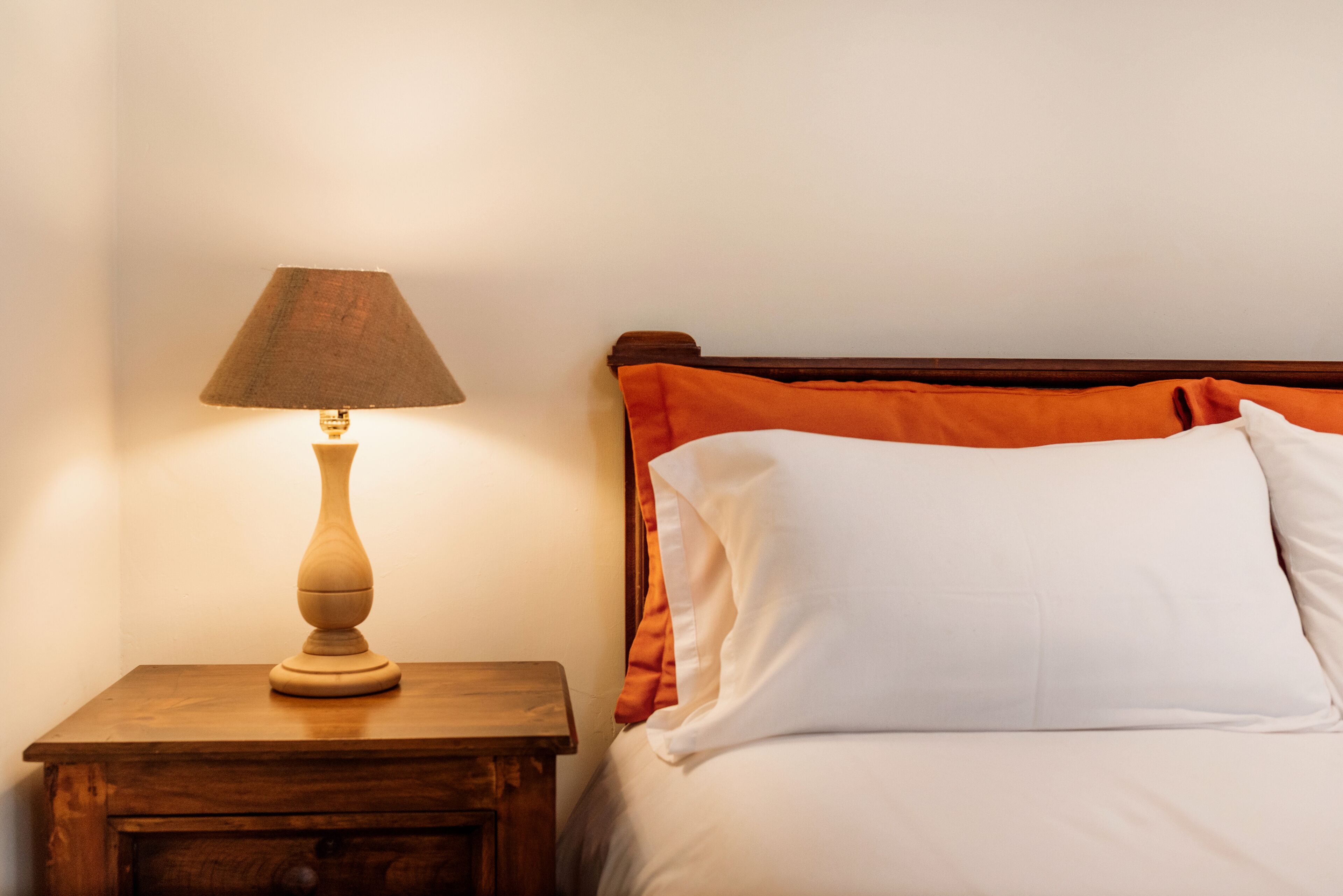 A wooden nightstand with a lamp, a bed with white and orange bedding, and a plain wall.