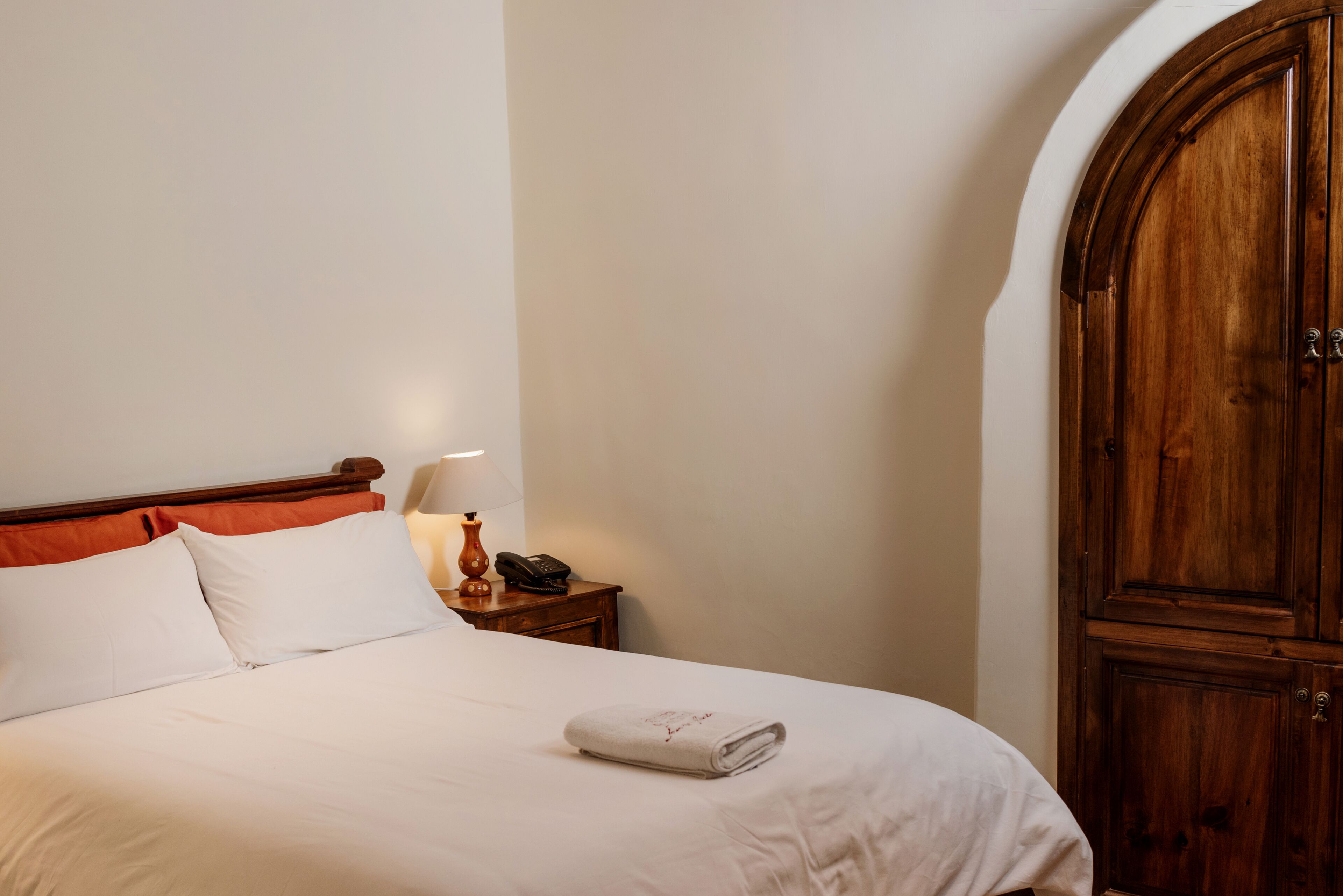 A neatly made bed with white linens and a folded towel, a bedside lamp, a telephone, and a wooden wardrobe in a hotel room.