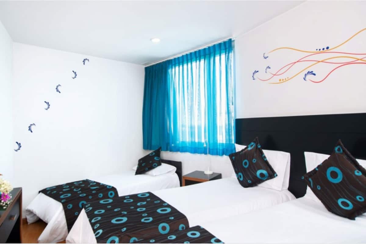 A hotel room with two beds, a large window with blue curtains, a wall with abstract art, and a floral arrangement on the nightstand.