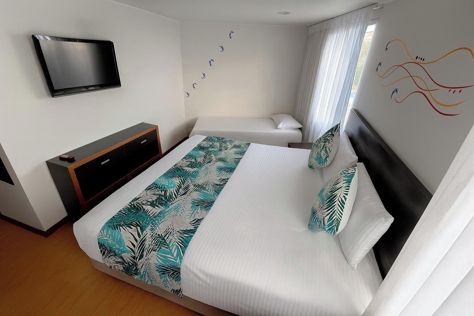 A hotel room with a bed, a TV mounted on the wall, a bedside table, and a window with curtains.