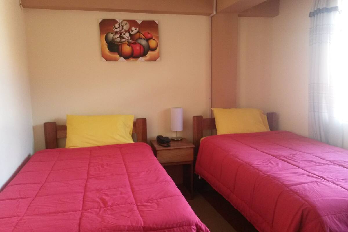 Habitación de hotel con dos camas, cada una con cubrecama rojo y almohada amarilla. En la pared sobre las camas hay un cuadro, una mesita de noche con lámpara y una ventana con cortinas.