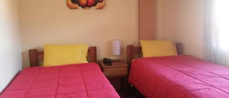 Habitación de hotel con dos camas, cada una con cubrecama rojo y almohada amarilla. En la pared sobre las camas hay un cuadro, una mesita de noche con lámpara y una ventana con cortinas.
