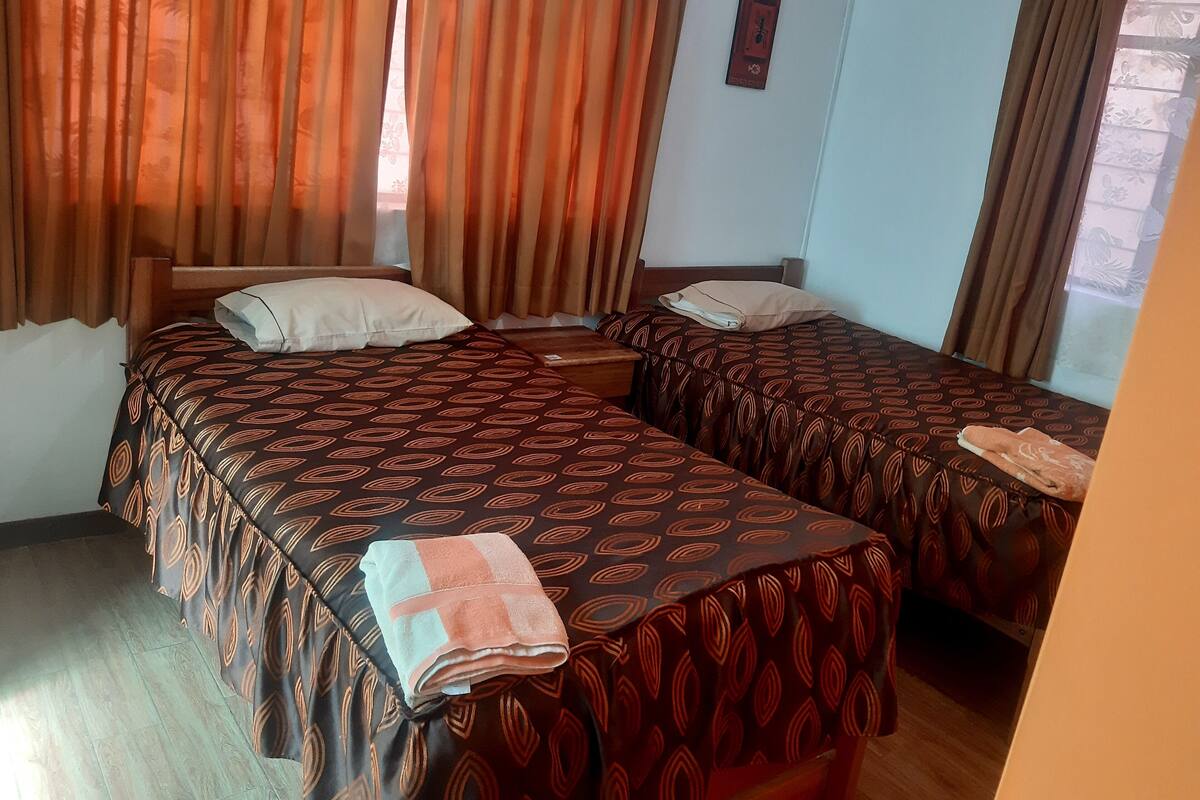 Habitación de hotel con dos camas, cada una con ropa de cama y almohadas estampadas, piso de madera y cortinas.
