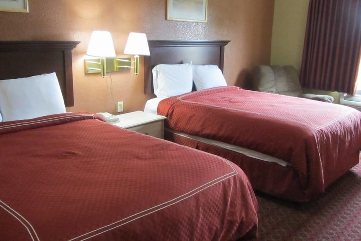 A hotel room with two beds, a nightstand, a lamp, and a window with curtains.